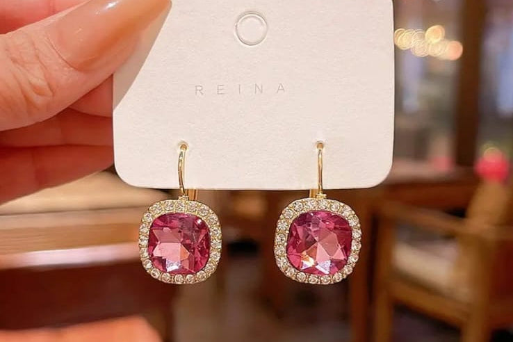 Pink gemstone earrings on a white card held by a hand with a blurred indoor background.